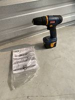 Ryobi HP496 cordless drill standing upright on a surface with instruction manual in plastic bag beside it. Shows general appearance and accessories included.