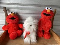 Set of three plush toys placed on a wooden chair: two Elmo dolls and one white dog plush with a red ribbon.