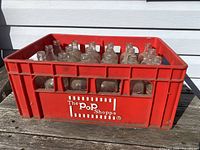 Red plastic crate marked 'The Pop Shoppe' holding clear glass bottles with red labels as viewed from the front outside.