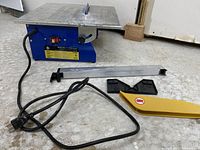 Photo of the blue portable tile saw on a floor with attached power cord and accessories (metal guide rail, clamps, yellow safety guard) arranged in front of it.