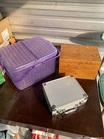Photo showing a purple plastic basket with lid, a natural wood box, and a silver metal case placed on a dark surface.
