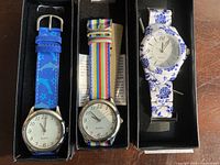Front view of three unworn analog quartz watches displayed in black boxes with unique colorful straps: blue star print, multicolor stripe, and blue floral.