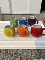 Set of six round, glossy ceramic mugs in vibrant colors: yellow, orange, red in front row; green, blue, purple in back row.