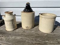Three vintage ceramic jugs on wooden surface against white siding: a tall jug with brown top and cork stopper, a milk jug with 'MILK' embossing, and a plain wide crock.