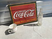 Wooden Coca-Cola sign along with glass bottle, ceramic mug, and glass on a flat surface with metal background.