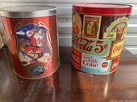 Photo of two large vintage Coca-Cola metal tins: one showing Santa Claus with silver lid and the other red lid with vintage ads.