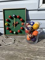Photo shows square pool ball clock with green felt background and red knobs as hour markers along with four numbered pool balls (3,6,9,12) and a clear glass container filled with colorful vintage pool balls on weathered wood surface outdoors.