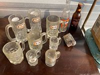 Set of 11 vintage glass items consisting of mugs and bottles from A&W, Hires, and Stewart's. Various sizes of mugs with logos, two glass bottles visible, one clear and one brown.