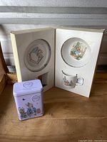 Full set including the open box with cutout slots holding the plate and cup, and the metal tin in front of the box.