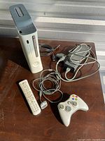 Photo of the Xbox 360 white console standing upright, with controller, remote, power brick, and AV cable on a wooden surface in natural light.