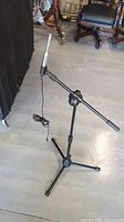 Microphone stand with tripod base and boom arm shown standing on light-colored floor with office chairs in background. Cable hanging from boom arm.