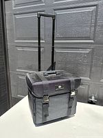 Closed black nylon makeup carrying case with telescoping handle extended and two silver latch clasps securing top lid against a dark garage door.