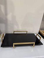Front view of rectangular black acrylic tray with four gold metal handles, showing glossy surface and handle design.