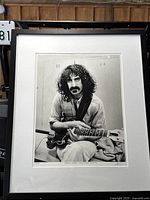 Framed black and white photo of Frank Zappa seated with guitar, showing the full frame and matting against a wood background.