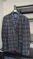 Grey men's blazer with a windowpane plaid pattern hanging on a black hanger. Shows front side with button closure and lapel details.