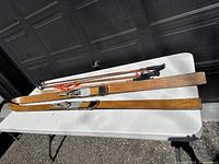 Wooden skis and four ski poles laid on a white folding table outdoors, showing overall length and shape of skis and poles.