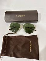 Oliver Peoples sunglasses with green lenses lying in front of the branded hard case and soft pouch on white background, showing front view of glasses and logos on case and pouch.