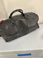 Front view of black leather duffel bag with two handles and red zipper puller, showing textured leather.