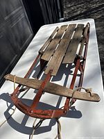Photo of vintage wooden sled with red metal runners and pull rope on white surface.