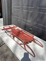 Vintage wooden sled with red painted metal frame showing some rust and wear, view of top and side surfaces.