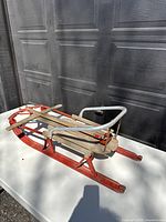 Side view of the vintage wooden snow sled showing the red-painted wooden runners, natural wood slats seat, and silver metal handlebar.