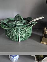 Green cabbage-shaped ceramic tureen with lid and white ladle on shelf.