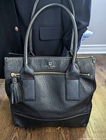 Front view of the black pebbled leather Kate Spade tote bag showing handle, front zipper pocket with tassel, and gold Kate Spade logo.
