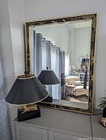 Front angled view of the mirror leaning on a console, showing black lacquer frame with gold chinoiserie floral design and reflective glass.