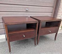 Front view of the pair of dark finished wooden oversized night tables with open shelf and drawer, placed on pavement in front of a garage door.