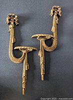 Two gold composite wall display shelves with bow and tassel motifs shown front side mounted on wall.