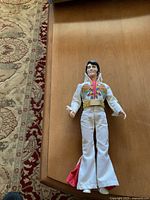 Full view of approximately 12 inch Elvis doll laying on wood surface, showing white jumpsuit with detailed embroidery and gold belt.