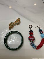 Photo shows a green bangle bracelet, a red and blue beaded bracelet with toggle clasp, and a multicolor beaded brooch with bohemian style design