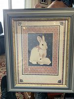 Frontal photo showing framed artwork of two white rabbits, the frame in blue with gold inner border and decorative patterned matting inside.