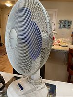 Front and side view of the Homestyles table fan showing blue blades behind white metal mesh cage, circular white plastic base with 4 blue buttons and white power cord coiled on base, placed on a kitchen counter.