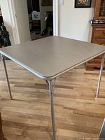 Beige square card table with metallic legs standing on hardwood floor, showing overall condition.