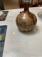 Glass round bottle with vintage map design and vertical real leather strips on a white table near chairs.