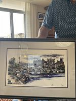Full view of framed Paris landscape art print with river, bridge, historic buildings and trees. Measures approximately 24 by 18 inches and includes wooden frame and matting.