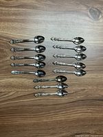 Top view of 13 silver-plated teaspoons arranged in two rows on wooden table showing decorative handles and polished bowls.
