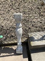 Full view of one decorative cement railing post placed outdoors next to a fence and steps.