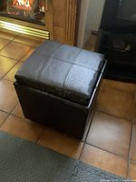 Dark brown square ottoman next to fireplace, top is removable and surface is worn.
