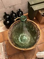 Top view of a large green glass demijohn with wicker basket around the bottom and sides alongside four smaller dark-colored glass demijohns in the background.