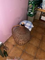 Large demijohn on tiled floor next to a small chair covered with a blue-and-white checkered cloth. Wicker cover and handle visible, sealed with cork.