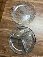Top view of two clear glass serving dishes on a wooden surface, one with floral etching and scalloped edges, the other divided into three sections with etched pine branch design.