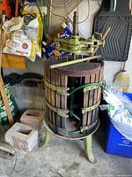 Full view of grape press showing wooden slats, metal bands, green metal base with legs, and attached hand crank.