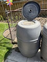 Large gray plastic barrel with removable black lid, positioned near a chain link fence in a grassy outdoor area.
