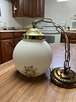 Photo of electric ceiling light fixture on wooden table in kitchen setting showing frosted glass globe with floral decoration and brass chain and ceiling mount.