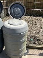 Gray cylindrical plastic rain barrel with removable lid, positioned outside near a chain-link fence.