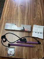 Portable Pilates resistance bar with attached cords, instruction booklet, and packaging box on wood surface.