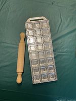 Photo showing entire aluminum ravioli tray with 24 square compartments plus wooden rolling pin on green cloth.