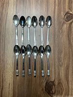12 silver-plated teaspoons laid out on wooden surface, showing front side with embossed decorative details on handles.